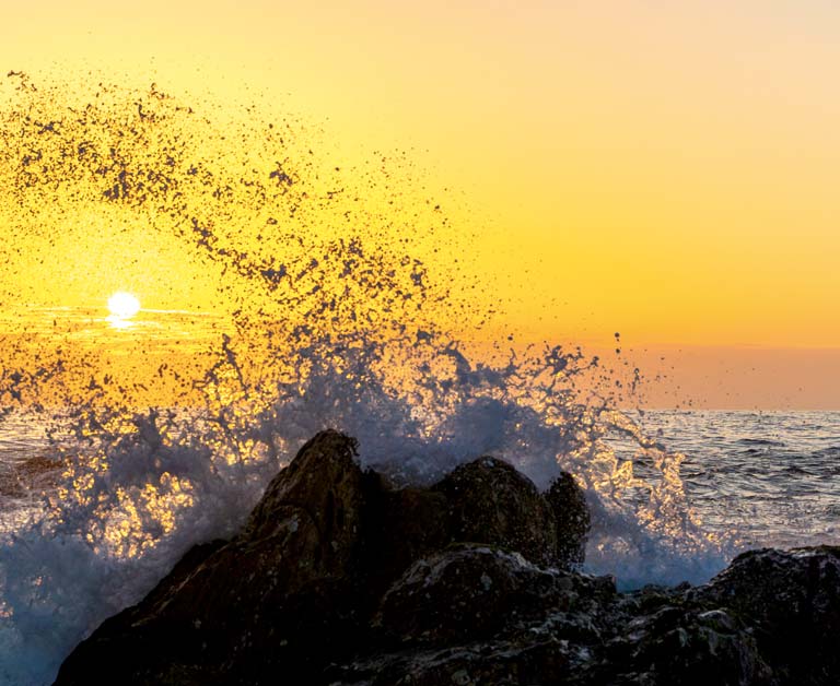 Ocean waves crashing against rocks in the foreground with sunset in the background