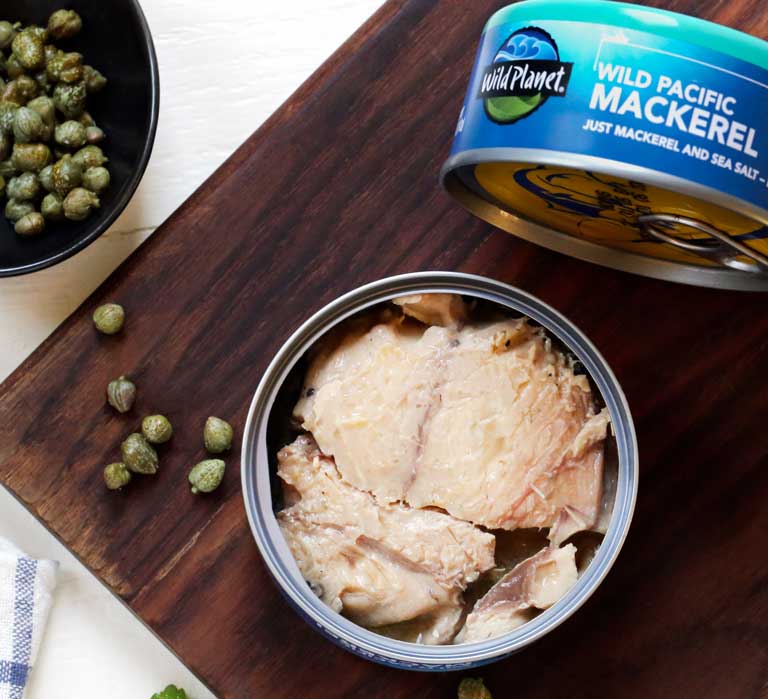 Cans of Wild Planet Wild Sardines in Extra Virgin Olive Oil, one open with a fork inside, one unopened, on a cutting board with fresh ingredients