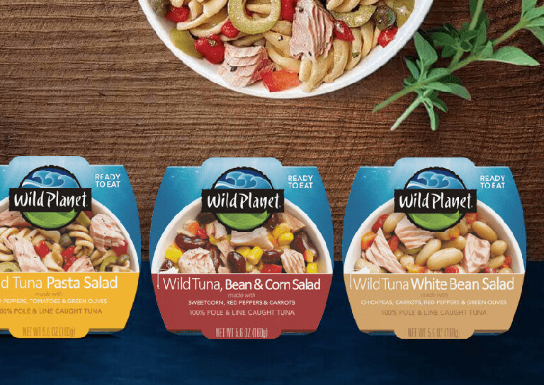 3 Wild Planet Ready to Eat Package: Wild Tuna Pasta Salad, Wild Tuna, Bean and Corn Salad, Wild Tuna White Bean Salad on a Table with a partial view of Salad Plate and side Cilantro